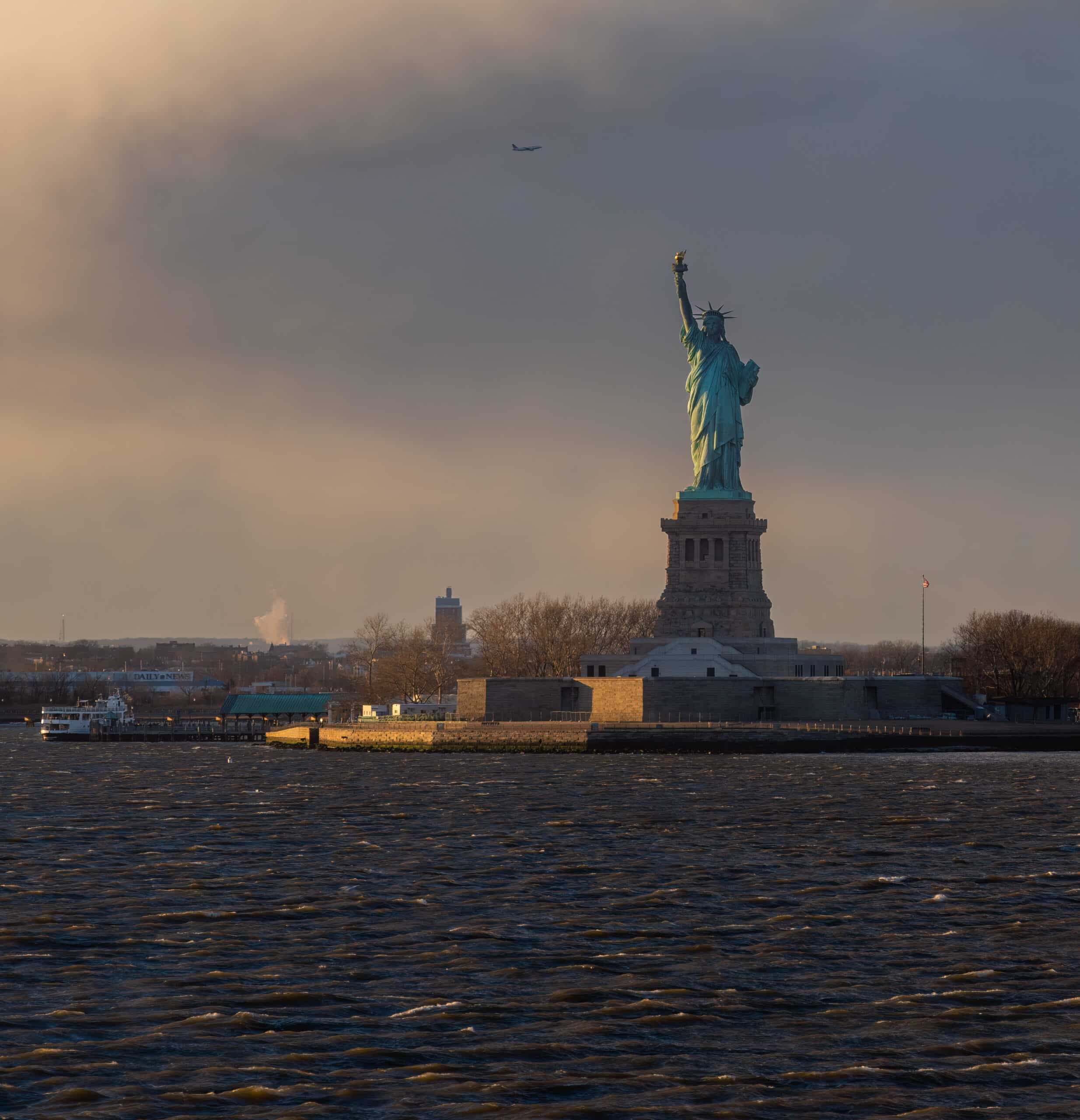 View of the Statue of Liberty during the day with boat docked on the island and Plane flying overhead